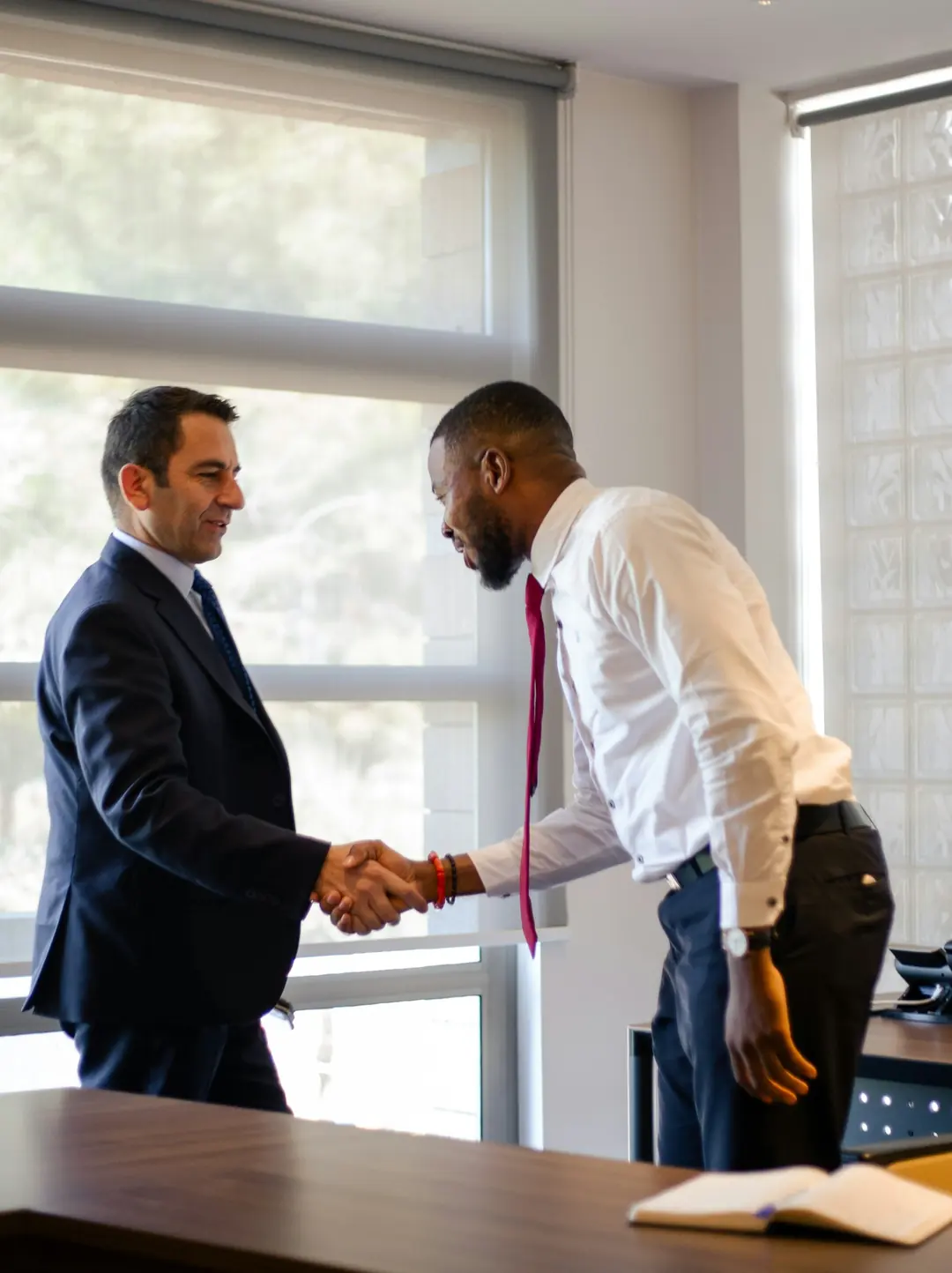 Dois homens sorrindo e apertando as mãos em um escritório claro, um deles de terno